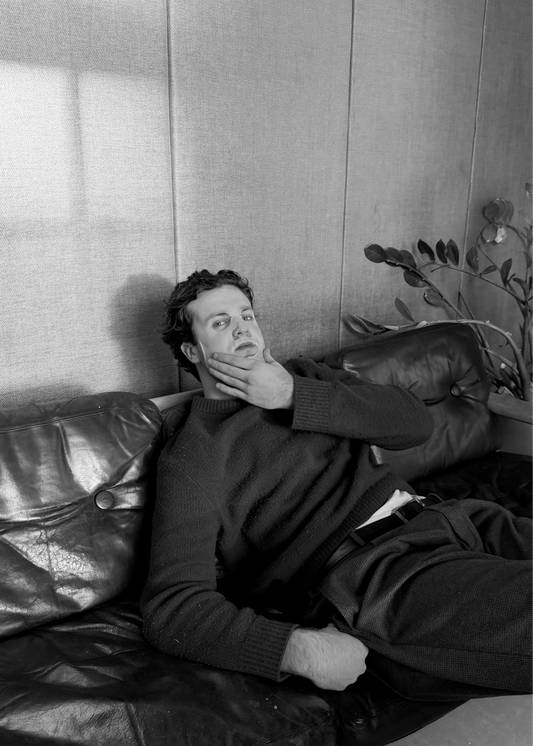Man lying on a leather couch with a sheet mask on in a room with a window and plant.