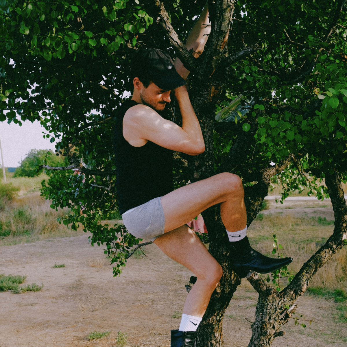 A man wearing a black tank top, gray shorts, white socks, and black boots is climbing a tree outdoors on a dirt path.