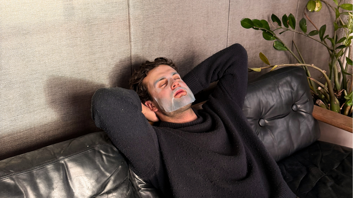 A man with a beard and a sheet mask on lies back on a black leather sofa against a textured beige wall, wearing a dark charcoal knit sweater and brown pants.