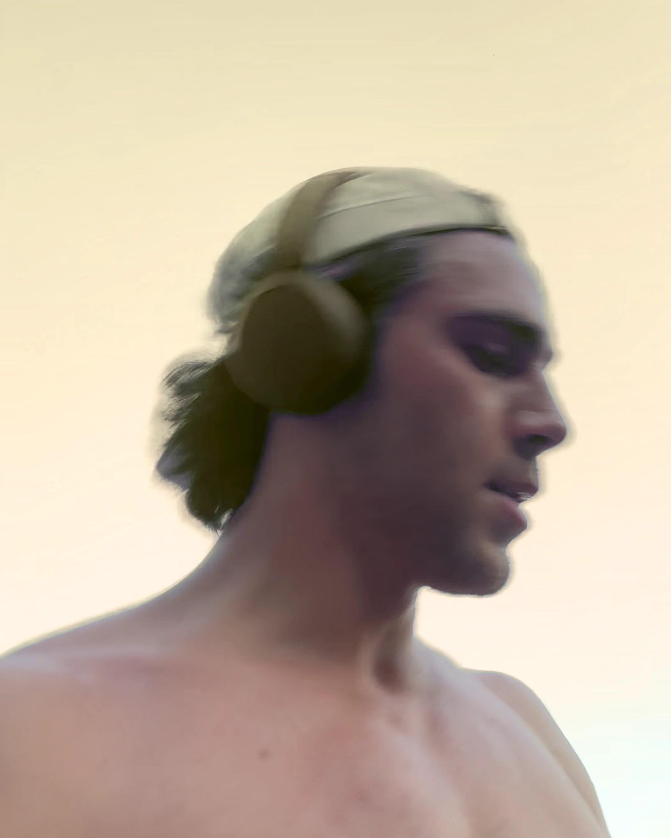 A close-up side profile of a shirtless man wearing over-ear headphones and a light-colored head covering against a plain light background.