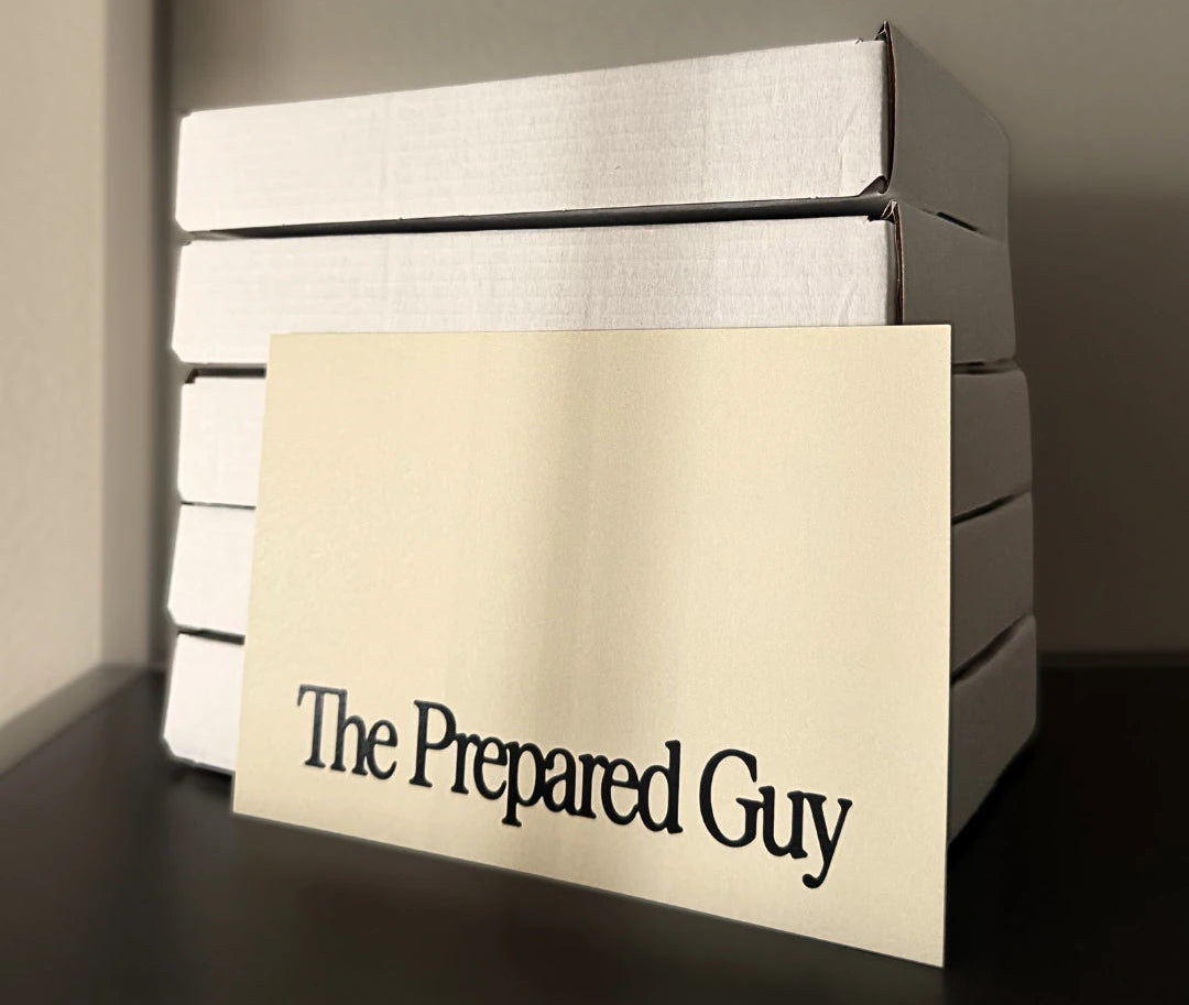 A cream-colored greeting card with the text 'The Prepared Guy' in black serif font, leaning in front of a stack of white boxes on a dark shelf against a neutral gray background.