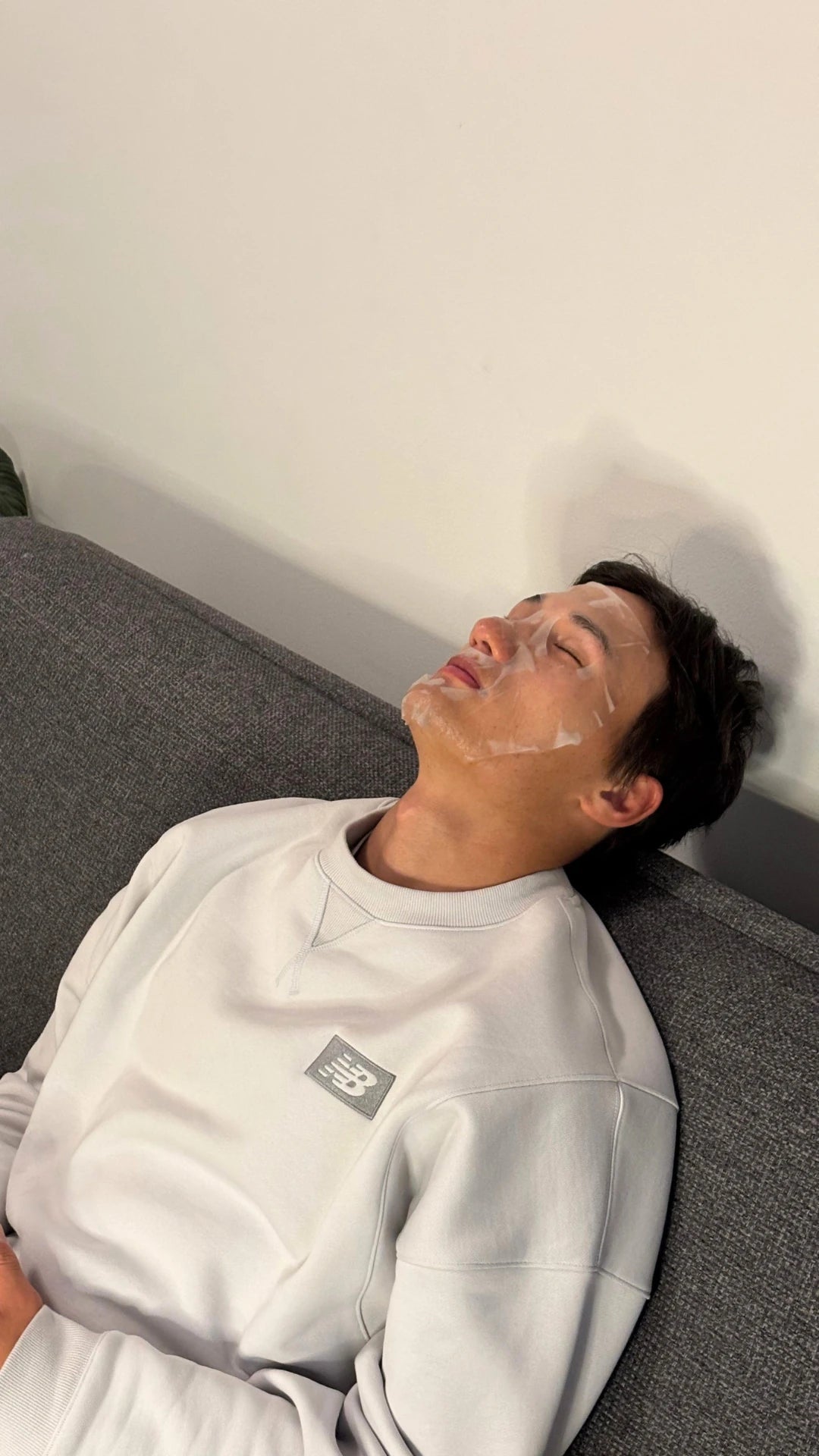 A man lying down with a sheet mask on, relaxing on a gray couch, wearing a light gray sweatshirt with a small square logo patch on the left chest.