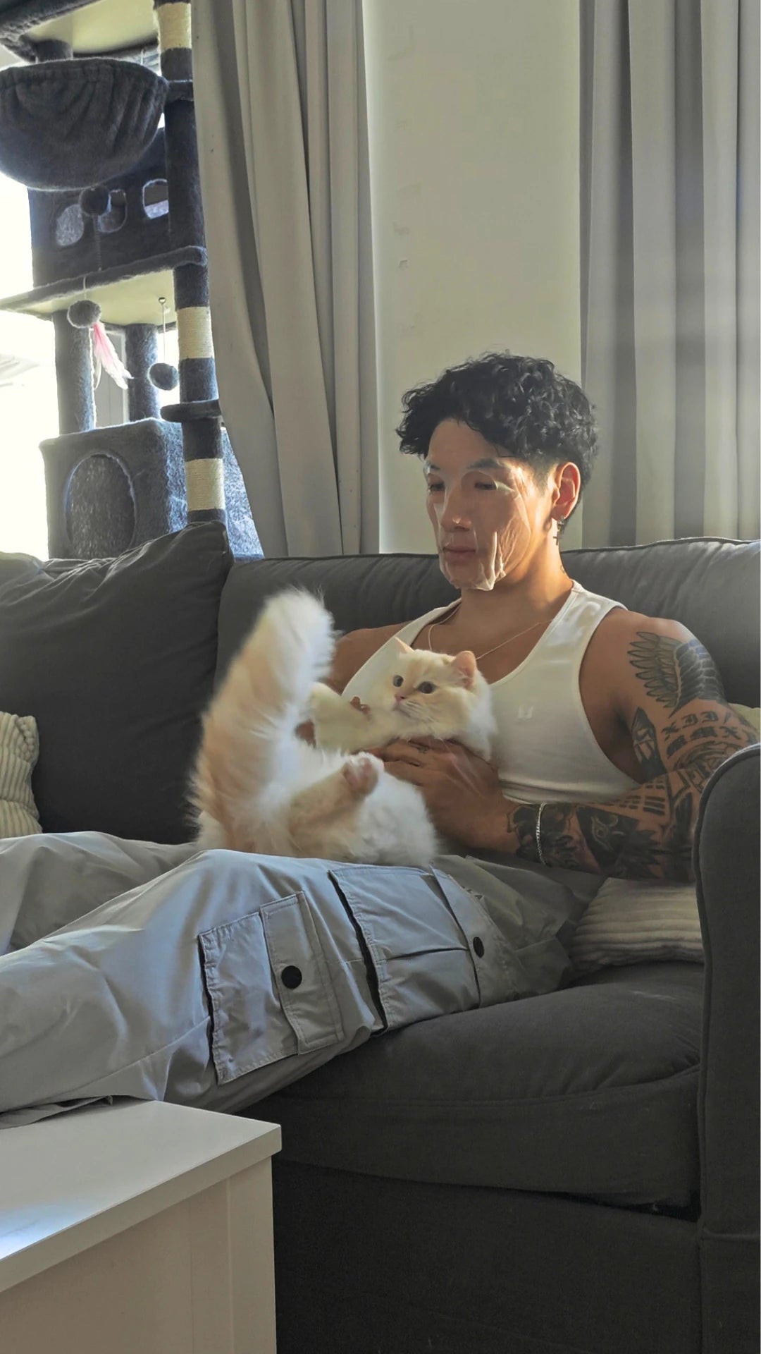 A man with a sheet mask on, has tattoos sits on a dark gray couch in a living room, holding a fluffy white cat. He is wearing a white tank top, light gray cargo pants, and has a cat tree visible in the background.