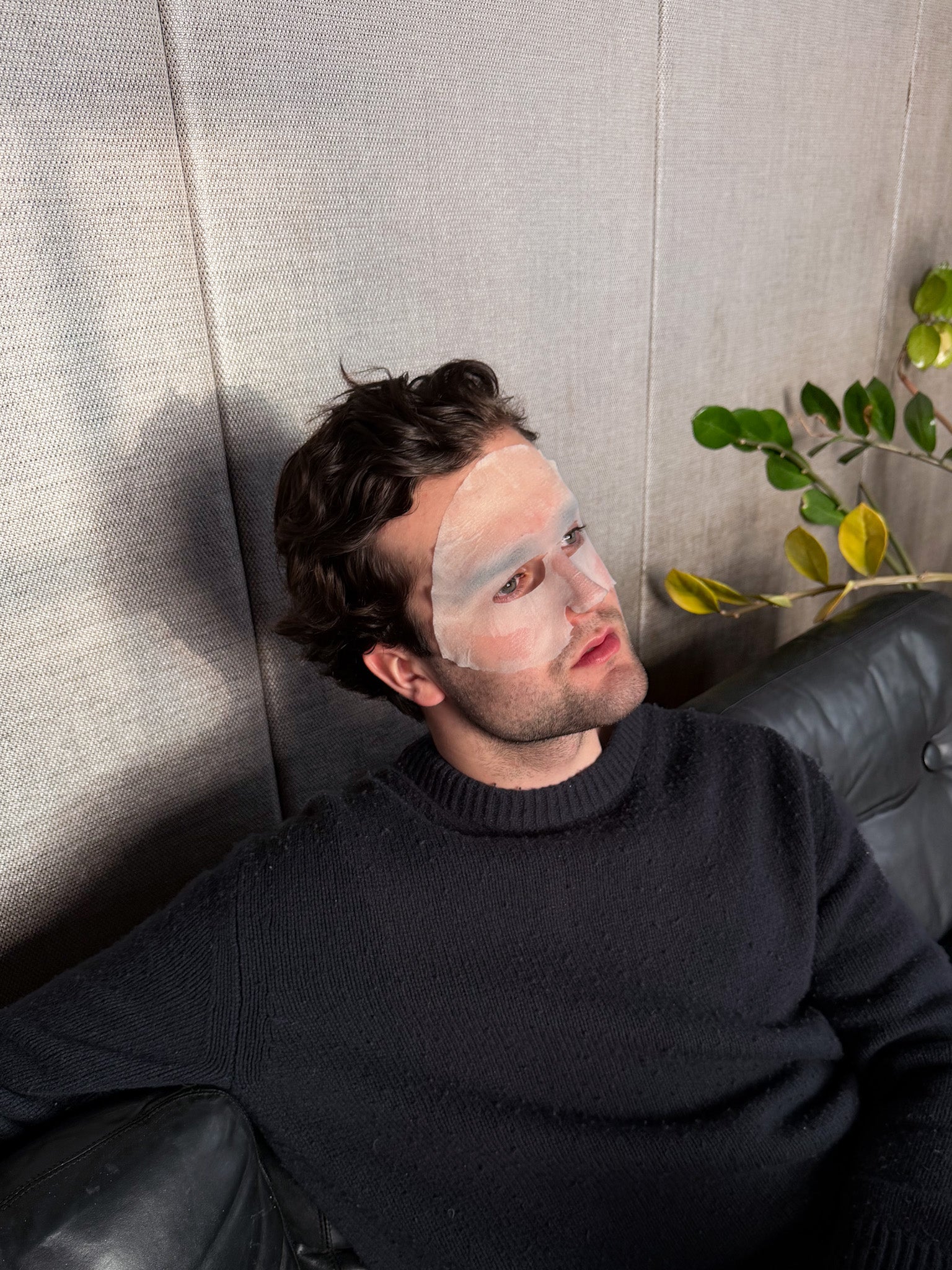 Man lying on a couch with a sheet mask, wearing a dark sweater.